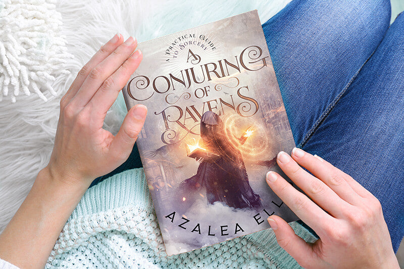 Person holding a Paperback of A Conjuring of Ravens